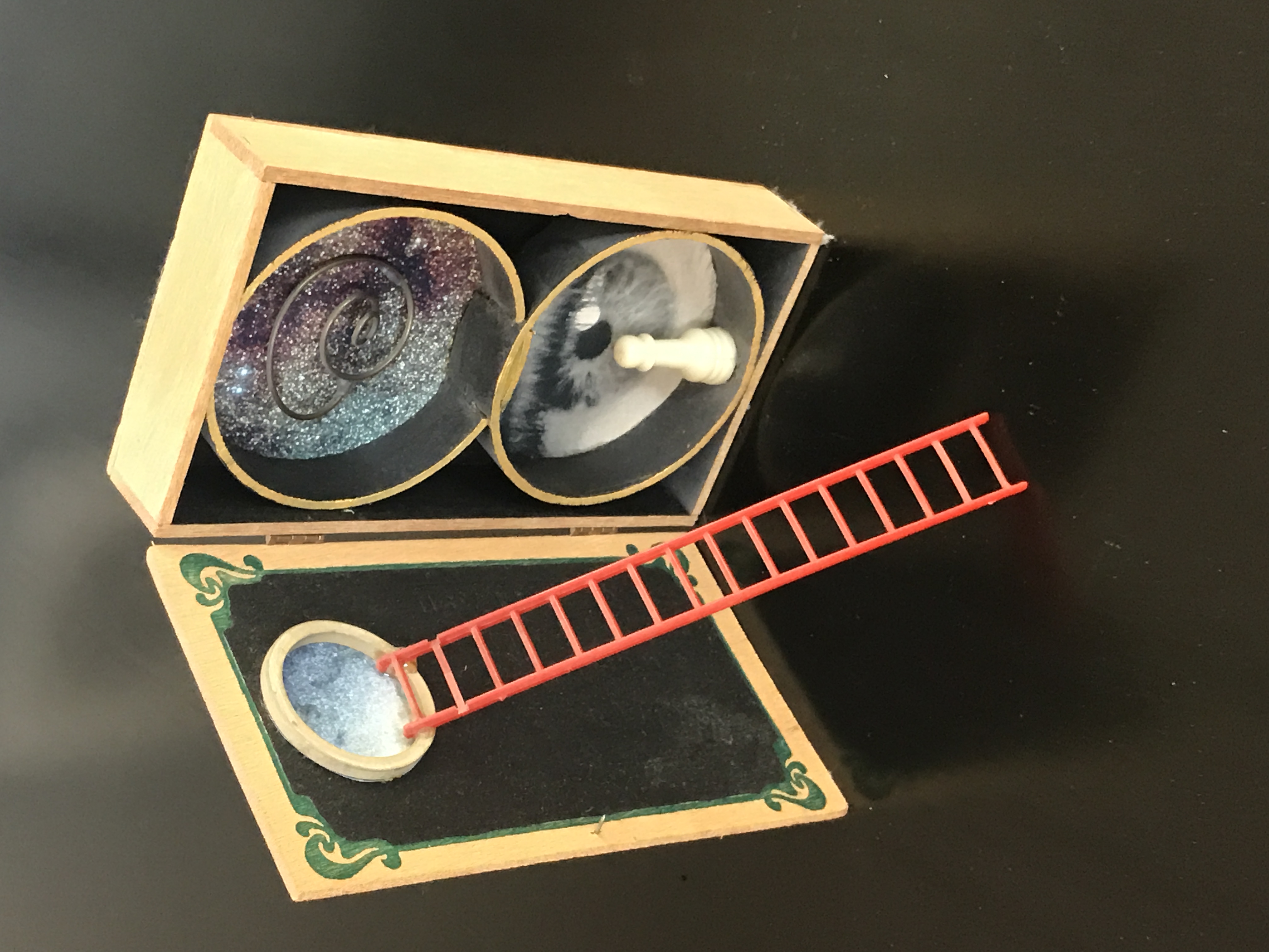 A box containing found objects assembled into a composition, with two circles on one side of the box, each containing a composition, and on the other side, a circle looks like a portal with a ladder leaning up against it.